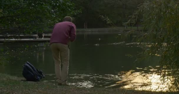 Mann kommt ans Ufer des Sees Mann mit Rucksack legt seinen Rucksack ab und sitzt am Wasser berührt das Wasser mit der Hand wirft den Stein ins Wasser — Stockvideo