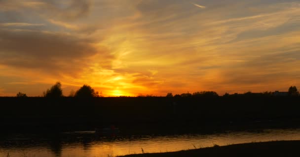 La barca galleggia vicino alla superficie dell'acqua nelle tenebre L'acqua scorre Tramonto giallo brillante al fiume Nuvole colorate Le silhouette degli alberi di cirro di fronte alla riva — Video Stock