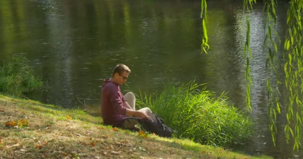 Ο άνθρωπος σε γυαλιά έρχεται στην όχθη του ποταμού κάθεται παίρνει το laptop ανεξάρτητος προγραμματιστής κειμενογράφος σχεδιαστής — Αρχείο Βίντεο