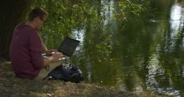 GözlükMan Slope Freelance Programcı Tasarımcı Copywriter Muhasebeci Oturuyor River Rippling Su Sırt Çantası Dizüstü ile Çalışıyor