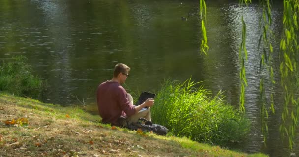 Man in Glasses está sentado en River Bank Programador independiente Diseñador de copywriter Accauntant está trabajando con el ordenador portátil Pone el portátil a la mochila se va — Vídeos de Stock