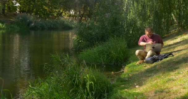 眼鏡をかけた男が川の斜面に座って、ラップトップフリーランスプログラマーデザイナーコピーライター会計士と一緒に働くラップトップを開いた — ストック動画