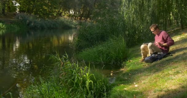メガネの男フリーランスプログラマーデザイナーコピーライター会計士は川の緑の斜面に横たわっている男は、ラップトップが仕事を始めるのに立ち上がった — ストック動画