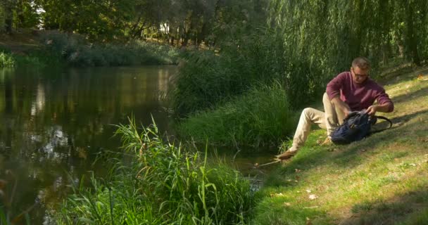 Homme dans les lunettes avec sac à dos programmeur indépendant concepteur rédacteur comptable vient à River Bank Slope travailler avec ordinateur portable fermé Posez-le vers le bas — Video