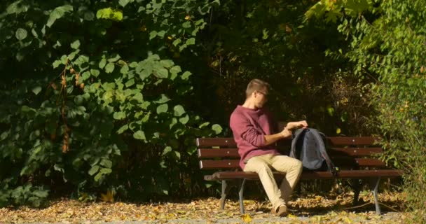 Man in Glasses Man With Backpack Comes to Bench Freelance Programmer Designer Copywriter Accountant Working with Opened Laptop Bushes are Behind — Stock Video