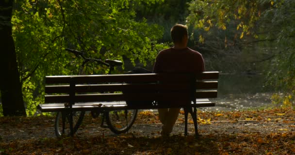 Man sitter på bänken mannens rygg cykel står nära man River vattenflöden man tittar på vattnet sitter med benen Pursed — Stockvideo