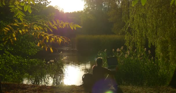 Silhouette de l'homme est assis sur la rive du lac avec ordinateur portable programmeur indépendant concepteur rédacteur comptable a tourné mettre l'ordinateur portable sac à main ses jambes — Video