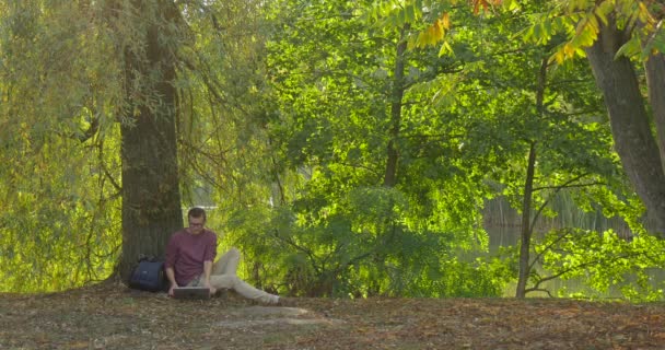 Uomo in occhiali è seduto sotto l'albero con il suo computer portatile messo via programmatore freelance progettista copywriter ragioniere sta meditando zaino sta mentendo — Video Stock