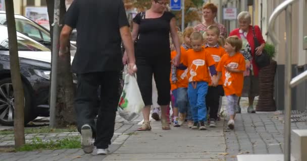 Nauczyciele prowadzą grupy dzieci w Orange T-shirty ludzie są Walking by City Street w kierunku Camera utwardzone Road City Street zaparkowane samochody — Wideo stockowe
