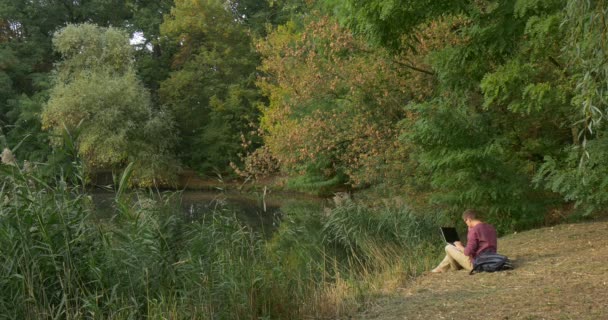 Man Sitting at The Lake Looking in Front of Him Man in Glasses Laptop está na mochila Freelancer Programador Copywriter Designer Contabilista Trabalhando — Vídeo de Stock