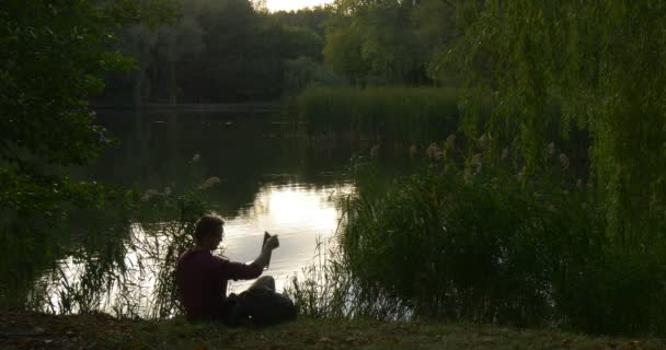 Человек сидит на The Lake Bank внештатный программист копирайтер дизайнер бухгалтер взял ноутбук работает глядя на экран ноутбука к озеру — стоковое видео