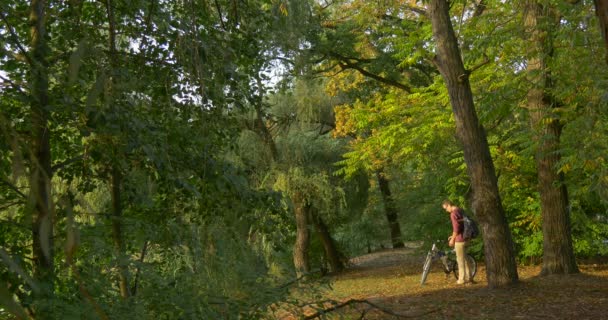 Чоловік приїжджає в ліс на велосипеді зняв свій рюкзак Сидить на землю зі своїм обличчям до променів сонця сидить під деревом — стокове відео