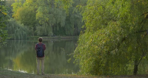Άνθρωπος σε γυαλιά με σακίδιο έρχεται να λίμνη Τράπεζα μόνιμης κοιτάζοντας τα ακουστικά του νερού είναι κρεμασμένα στο σακίδιο περπάτημα κάτω με νερό μετατρέπεται περπατά μακριά — Αρχείο Βίντεο