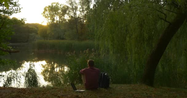 Homem está sentado no lago banco homem com Laptop Freelancer programador Copywriter Designer contador tem colocar seu Laptop olhando Sunset curta distância — Vídeo de Stock