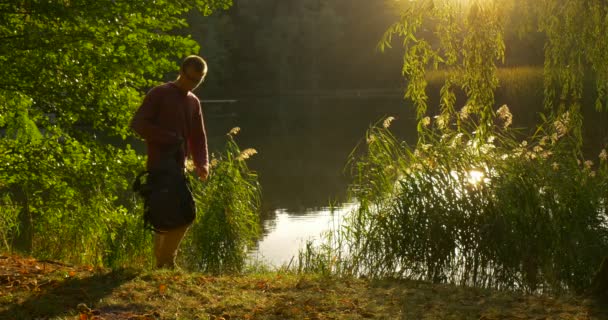 Man With Backpack is Standing With His Face to the Sun Rays Man at the Lake Bank Forest Sits to the Ground Ponga la mochila Lake Green Fresh Branches — Vídeos de Stock