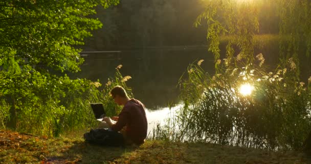 Mann sitzt am Ufer des Sees sitzt auf dem Boden mit Laptop Freelancer Programmierer Werbetexter Designer Buchhalter arbeitet den Laptop abgelegt — Stockvideo