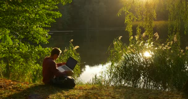 L'homme à lunettes est assis au lac avec son visage au soleil a pris l'ordinateur portable programmeur pigiste rédacteur concepteur comptable travaillant avec ordinateur portable — Video