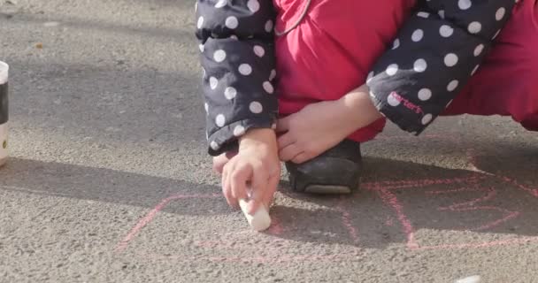 Niña está pintando a pie por tiza colorida Niña de las manos de la niña en chaqueta en lunares Niña en pantalones rosados Niña está sentada en el asfalto — Vídeo de stock