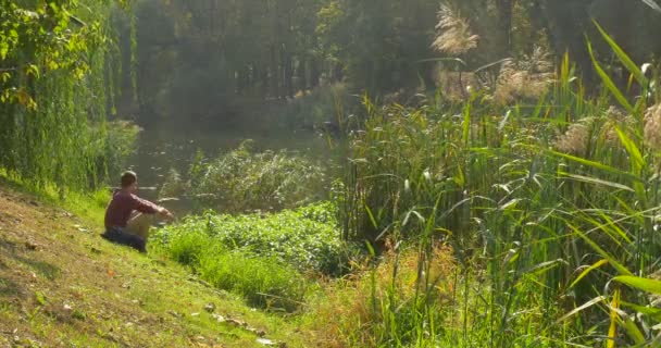 男子背着背包坐在草在倾斜的湖岸的人正在看水人转身脱下小书与棕色盖从背包 — 图库视频影像