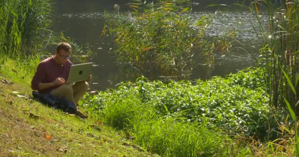 Hombre con gafas ha tomado el portátil de la mochila Hombre está sentado en el lago Banco Freelancer Programador Corywriter Diseñador Contador está trabajando — Vídeos de Stock