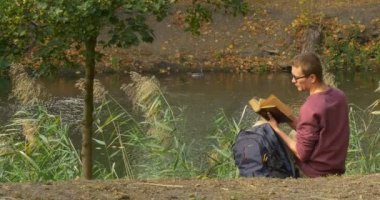 Gözlüklü Adam Yerde Oturuyor Adam Yiyor ve River Bank Kahverengi Kapak Adam Kitap Okuyor Sırt Çantası Içine Kitap koyun Away Walks