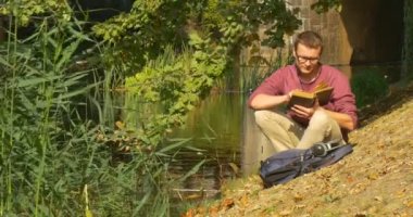 Gözlüklü Adam Suya Yakın Oturuyor Kitap Okuyucu kitabını kapattı Sırt Çantasına Koydu Yeşil Sazlık Rüzgar Güneşli'de Sallıyor