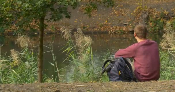 Człowiek w okularach siedzi na ziemi człowiek wziął książkę z plecaka człowiek czyta na brzegu rzeki Green Reed jest kołysząc Breeze — Wideo stockowe