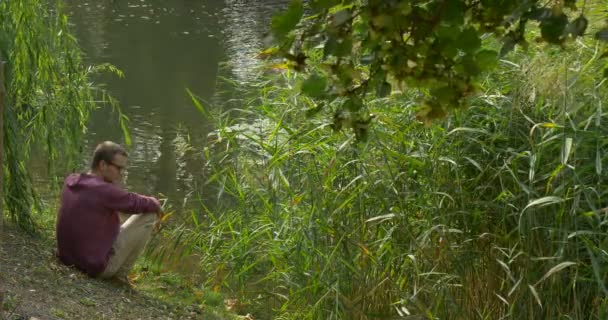 Az ember ül közel a víz és a buja zöld növények ül az ő lába Pursed és Arms keresztbe Man felállt elszalad a ferde Lake Bank zöld Reed — Stock videók
