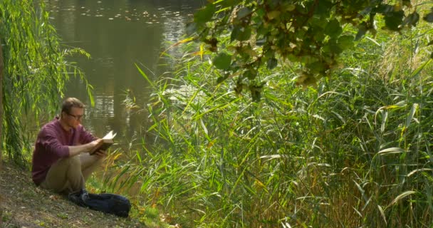 L'homme à lunettes est assis près de l'eau La lecture du livre Le lecteur feuillette les pages Assis sur le bord de la rivière en pente Sac à dos Green Reed — Video