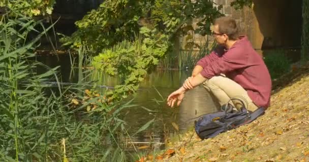 Muž v brýlích s batohem přichází ke svažující se bance u řeky sedí se kniha čtení knihy prolistování po stránkách slunečného dne podzim — Stock video