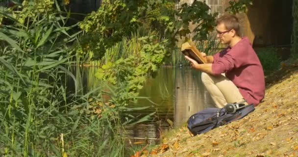 Man i glasögon sitter och läser boken om sluttande bank vid floden mannen tittar runt och tittar på fåglar gräsänder på vattnet solig dag — Stockvideo