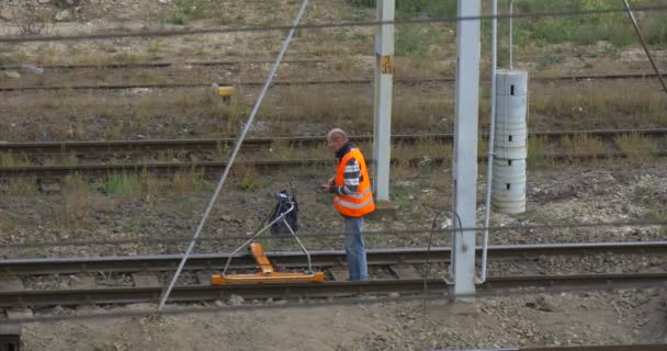 Hombre trabajador en ropa de trabajo naranja está caminando por el ferrocarril Comprobación del ferrocarril con una herramienta especial sobre las ruedas Hombre deja de caminar de nuevo Torre de alambre — Vídeos de Stock