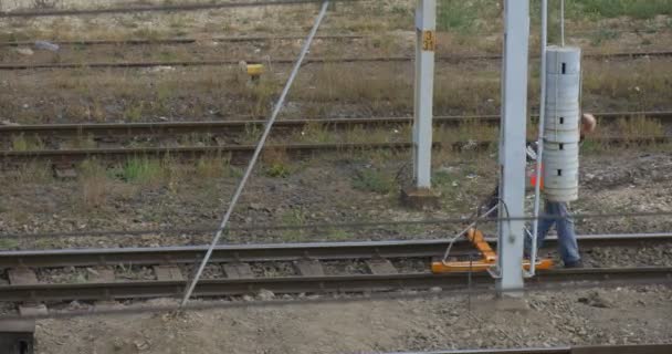 Hombre trabajador en ropa de trabajo naranja está caminando por el ferrocarril Comprobación del ferrocarril con una herramienta especial en las ruedas Torre de alambre a mediados de tiro al aire libre Nublado — Vídeo de stock