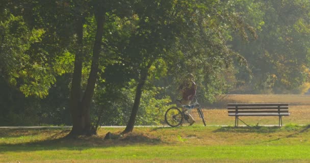 Mannen med ryggsäck står vid bänken mannens tillbaka mitten av skott cykel är ständiga Man fick på cykel vände och ridning bort vid vägen till parken — Stockvideo