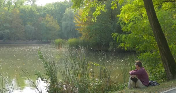 Man is Sitting at The Lake Bank in Forest With Book Looking at Water Takes the Laptop Freelancer Programmer Copywriter Designer Accountant Working — Stock Video