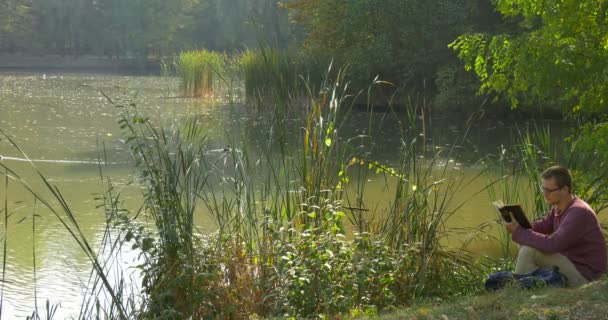 L'homme à lunettes est assis sur le sol sur le bord du lac dans la forêt et la lecture du livre Feuilleter les pages Regardant vers la droite Mettre le livre Quitter — Video