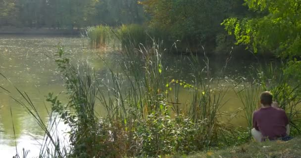 Людиною, в окулярах сидить за The Lake банку і читання книги кладе книгу в рюкзак дивлячись на воду покласти в рюкзак на і прогулянки звідси — стокове відео