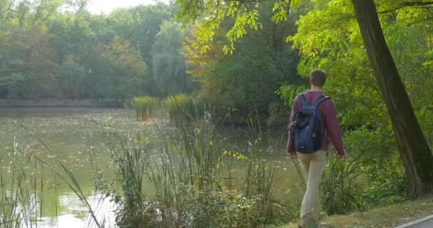 Людиною, в окулярах, з рюкзака сходить до озера банку в лісі взяти свій рюкзак і сидить на землю під зеленим акації, дивлячись на воді людина повернулася — стокове відео