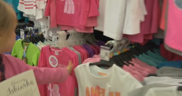 어린 소녀 t-셔츠와 선반을 따라 간다. 큰 상점에 의류 학과. — 비디오
