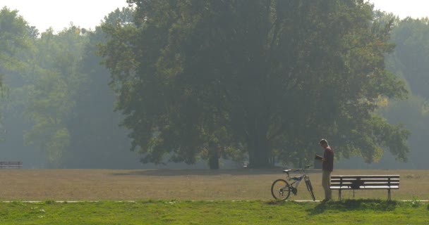 Mann holt Buch aus Rucksack, der im Park unterwegs ist, liest ein Buch Fahrrad steht an der Bank Mann fährt Fahrrad an sonnigem Tag im Hintergrund — Stockvideo