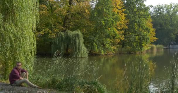 Człowiek w okularach siedzi na jeziorze Bank Man okazuje patrząc wstecz stoi i patrząc na Water Green Reed i gałęzie drzewa są kołysząc się na wiatr — Wideo stockowe