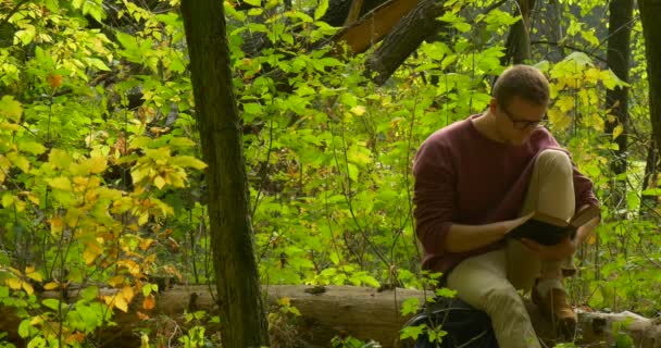 Uomo in Occhiali è seduto sul tronco dell'albero caduto e la lettura di un libro che l'uomo ha borsato la gamba sfogliando pagine chiuse l'uomo del libro ha girato — Video Stock