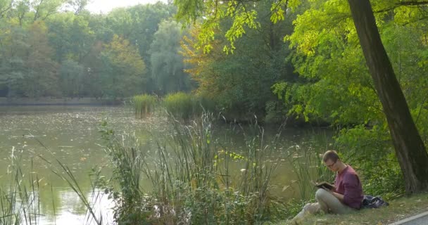 L'uomo è seduto alla riva del lago nella foresta con il libro Leggendo il libro che sfoglia le pagine L'uomo ha chiuso il libro Osservando la natura Guardando — Video Stock