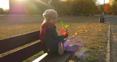 Küçük sarışın kız bere kırmızı eşarp ve ceketi giyen buket yaptı, yaprakları ve çiçekleri oturan bankta Park sokak binalar günbatımı içinde tutuyor