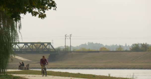 L'uomo in occhiali con lo zaino sta cavalcando la bicicletta verso la macchina fotografica che si muove dal vicolo asfaltato La gente è seduta al banco Le foglie secche stanno cadendo — Video Stock