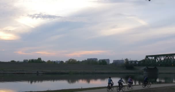 Cuatro personas cabalgan en las bicicletas en la orilla del puente del río a través del río Edificios residenciales Árboles verdes y hierba Cielo nublado Amanecer — Vídeos de Stock