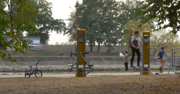 Esportes chão playground mulher faz exercícios duas meninas em patins pessoas andam com cachorro criança em bicicleta capacete estacionado azul bicicleta árvores verdes — Vídeo de Stock