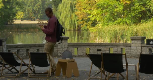 Человек с рюкзаком подходит к шезлонгу, снимает рюкзак и садится возле стола, человек берет чайник и наливает чай в зону чашечного парка озера — стоковое видео