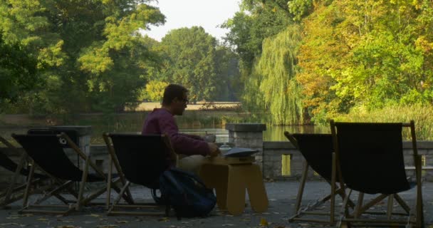 Man Pours Tea From The Teapot Man senta-se na cadeira de praia com xícara em suas mãos e olha para o lago vista para trás Park Area Lake Sunny Autumn Day — Vídeo de Stock