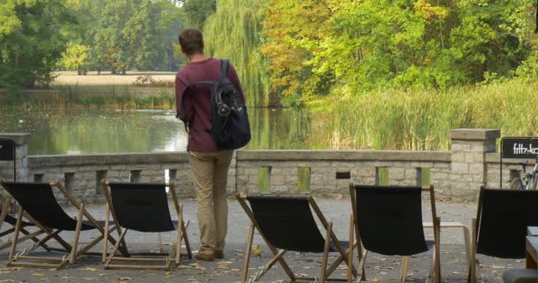 男は、彼は彼のバックパックを脱ぐと椅子に地上人の座っている上にそれを置くし、グリーン レイク公園エリア湖を見てデッキチェアの木 — ストック動画